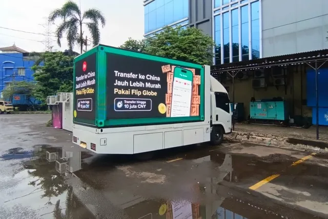 Truk LED putih dengan layar besar di sisi bak menayangkan iklan layanan transfer uang, terparkir di halaman